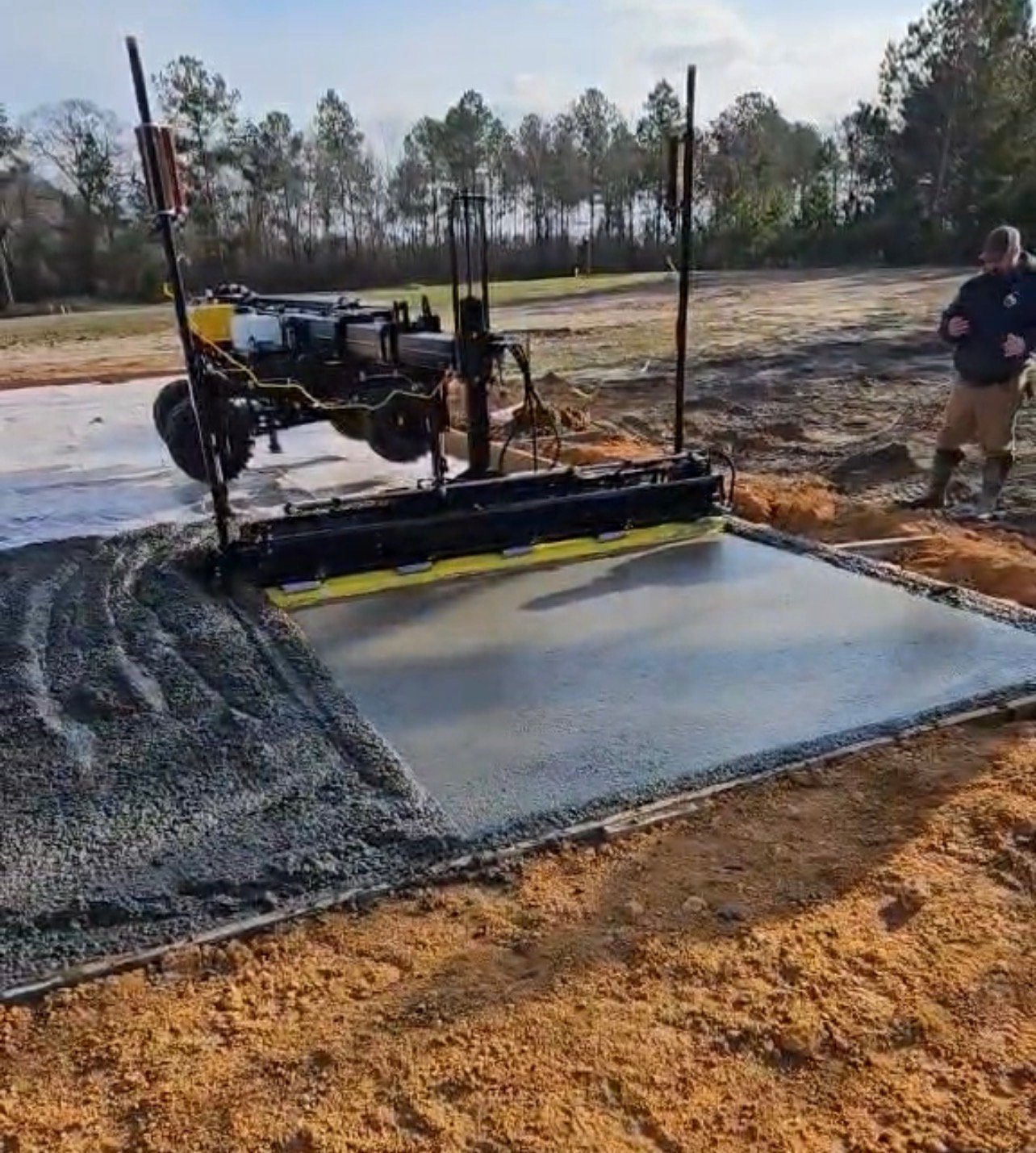 Asphalt paving machine laying hot tar on a concrete foundation at a construction site with workers and forested area in background