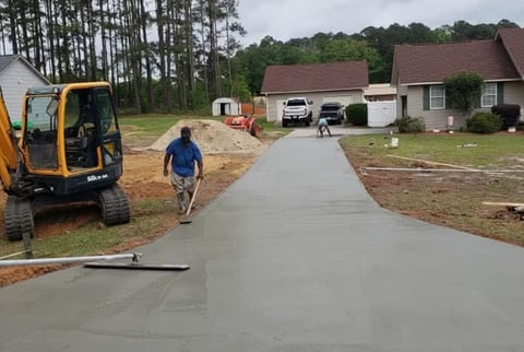 Workers pouring concrete driveway at residential home with yellow excavator and freshly laid cement
