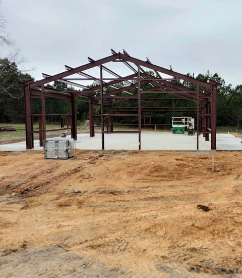 Reddish-brown metal building frame under construction on bare dirt ground with trees in background