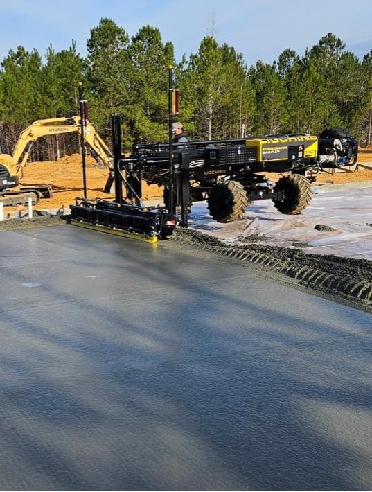 Road construction equipment spreading asphalt on a wet concrete surface with pine trees in background