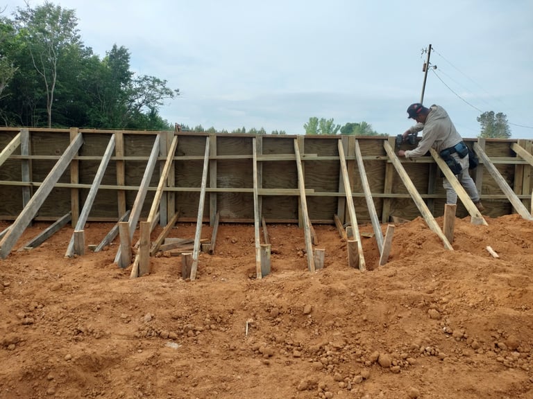 Worker on wooden roof framing structure under construction on bare earth with trees in background
