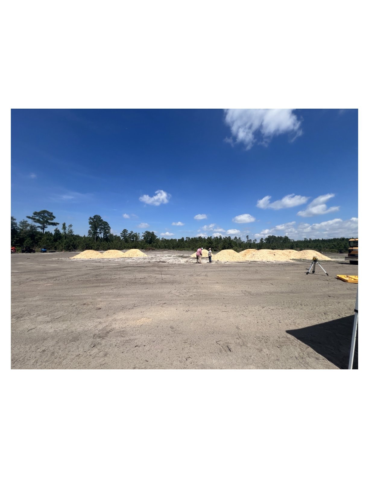 Large sandy construction site with piled materials, workers, and tree-lined horizon under blue sky with white clouds