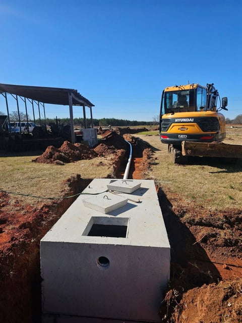 Hyundai excavator installing a white septic tank in a trench at a construction site with storage shed visible