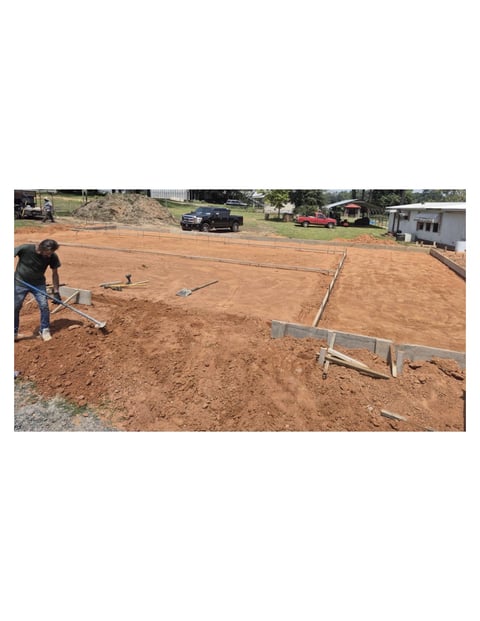Worker preparing reddish-brown soil foundation with string lines and metal guide, residential property in background