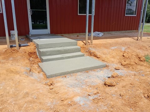 Concrete steps and walkway under construction leading to a red metal building on bare dirt ground