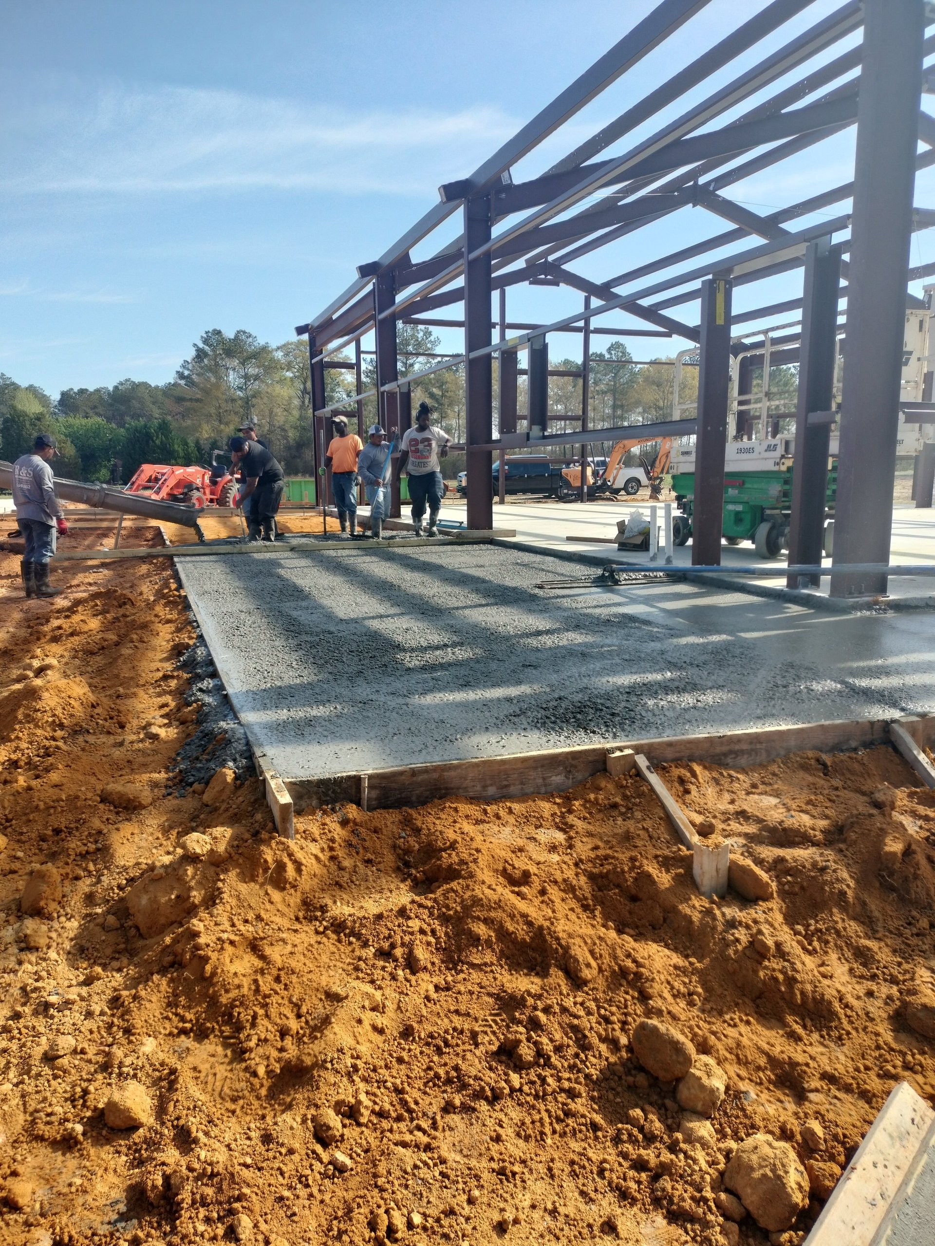 Construction site with concrete foundation and steel beam structure frame under bright sunlight, workers visible in background