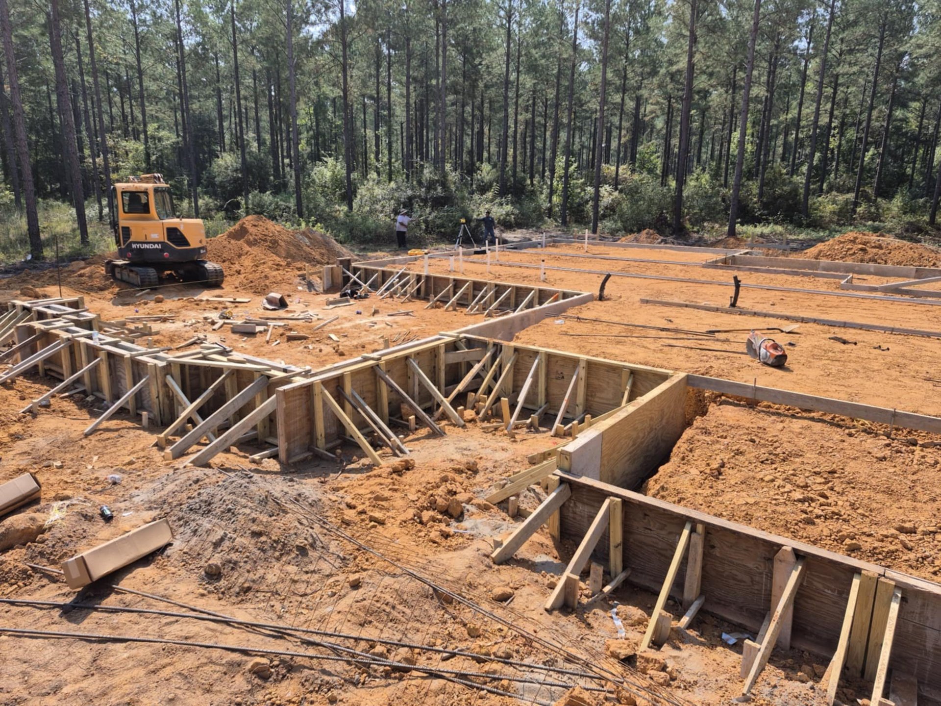Construction site with foundation framing and wooden supports, surrounded by pine forest and yellow excavator