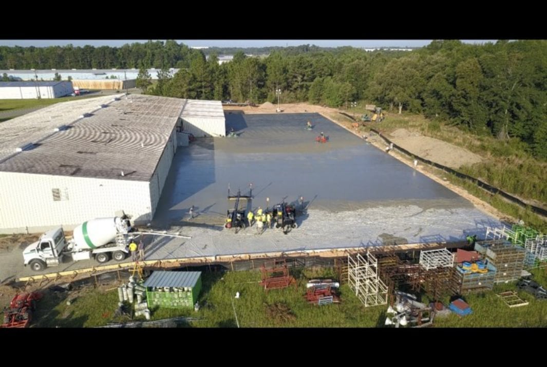 Aerial view of concrete pool deck under construction with workers, equipment, and surrounding industrial facility