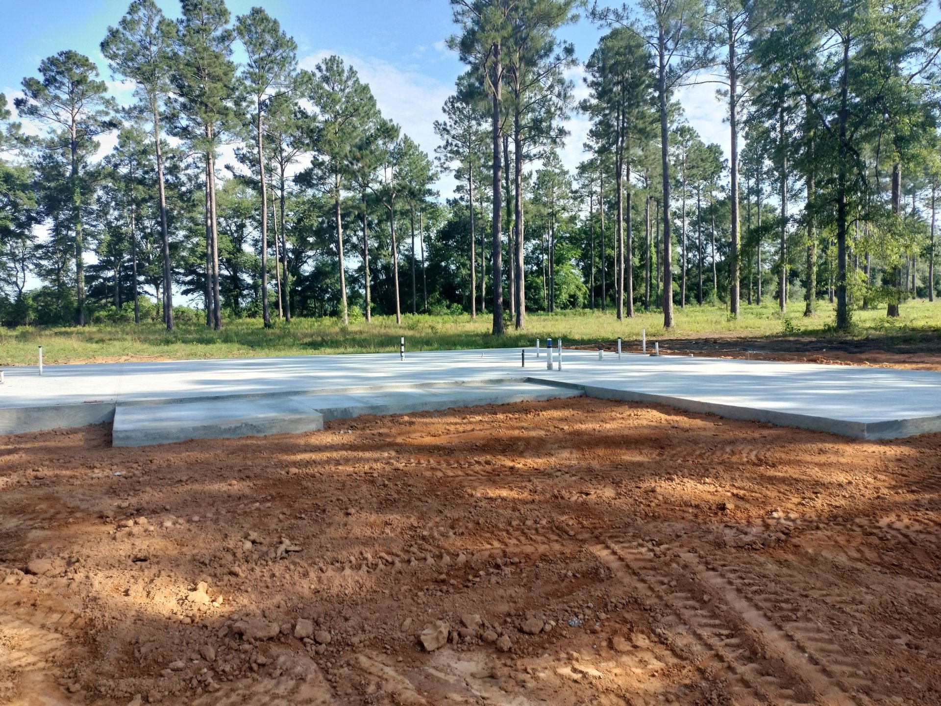 Construction site with concrete foundation slab laid on brown soil, tall pine trees in background, and clear sky