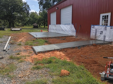 Construction of concrete foundation pads in front of a red metal building with trees and parked vehicles visible in the background