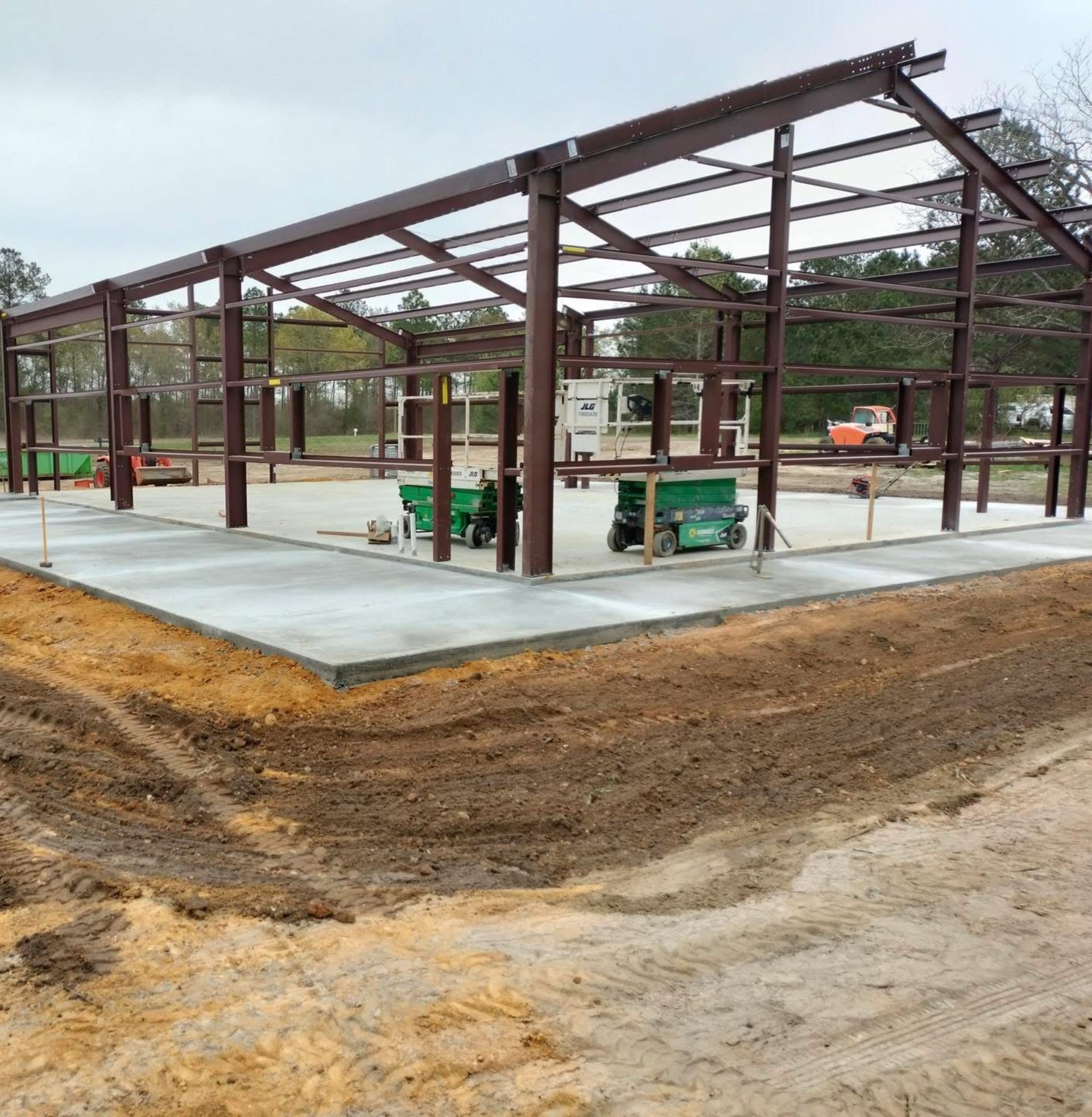 Construction site with brown metal frame structure on concrete foundation, surrounded by dirt and construction equipment in background