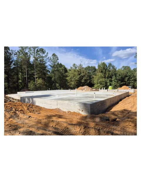 Construction site showing concrete foundation with white walls in cleared woodland area surrounded by excavated brown soil