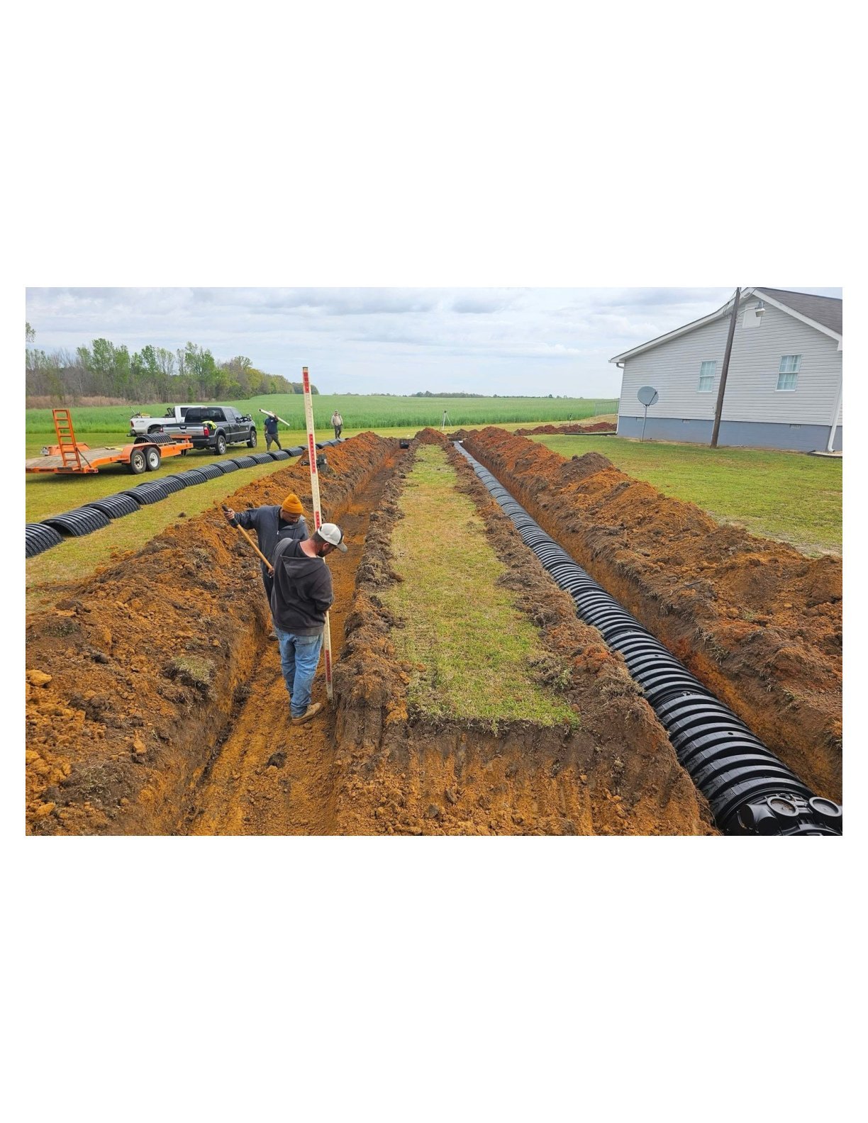 Worker installing black drainage pipes in trenches on rural farm property with equipment and house visible