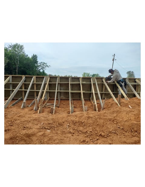 Worker constructing wooden roof frame on building foundation with exposed beams and trusses over bare ground